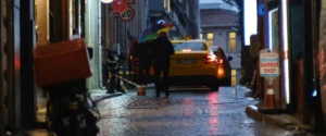 A person with a rainbow umbrella walks down a wet cobblestone street in Istanbul at dusk, past a yellow taxi and shop signs reflected in the rain